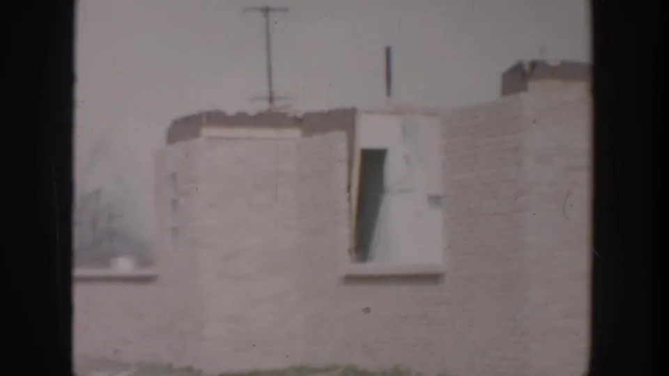 1956 Grand Rapids home-movie frame showing a pale brick wall, small window opening, roofline, and utility pole in the background.