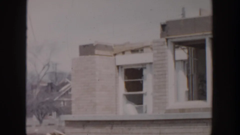 1956 Grand Rapids frame showing the brick building from another angle with window openings, bare branches, and adjacent construction details.