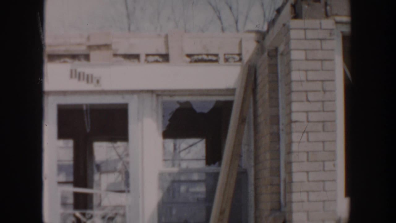 1956 Grand Rapids home-movie frame showing a damaged residential facade with broken window glass and adjacent brickwork.