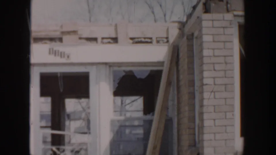 1956 Grand Rapids home-movie frame showing a damaged residential facade with broken window glass and adjacent brickwork.