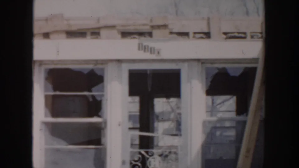 1956 Grand Rapids frame of a deteriorated house exterior with shattered panes, pale trim, and bare branches reflected in the glass.