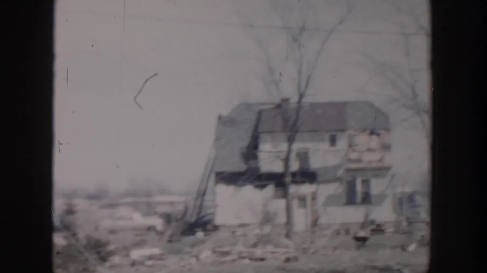 1956 Grand Rapids home-movie frame with a wider view of a damaged residential building, leafless trees, debris, and open ground.