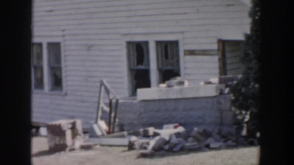 1956 Grand Rapids storm aftermath showing a white residential wall with broken windows, masonry, lumber, and scattered debris.