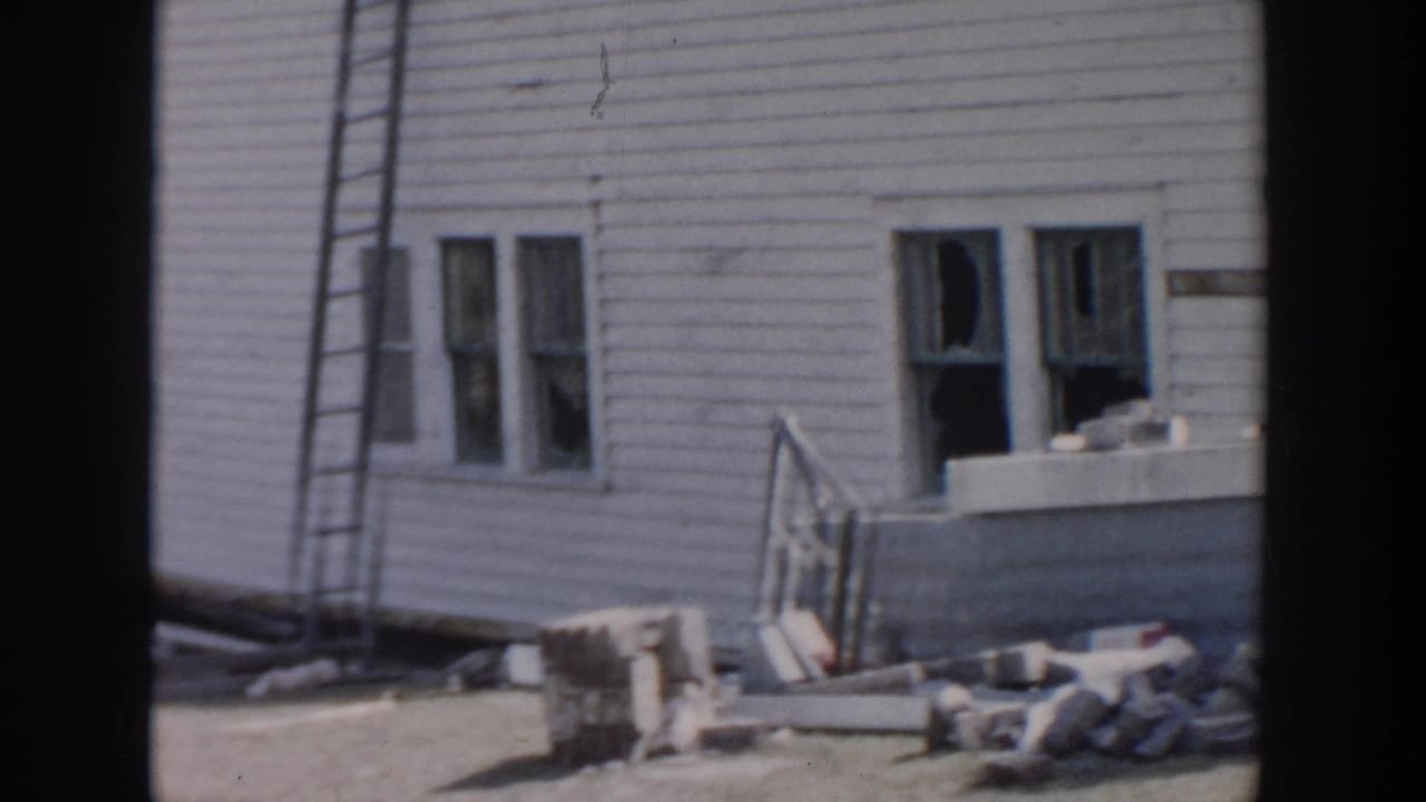 1956 Grand Rapids home-movie frame showing a damaged white clapboard house, broken windows, a ladder, and scattered debris.