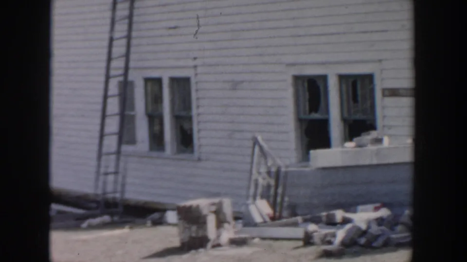 1956 Grand Rapids home-movie frame showing a damaged white clapboard house, broken windows, a ladder, and scattered debris.