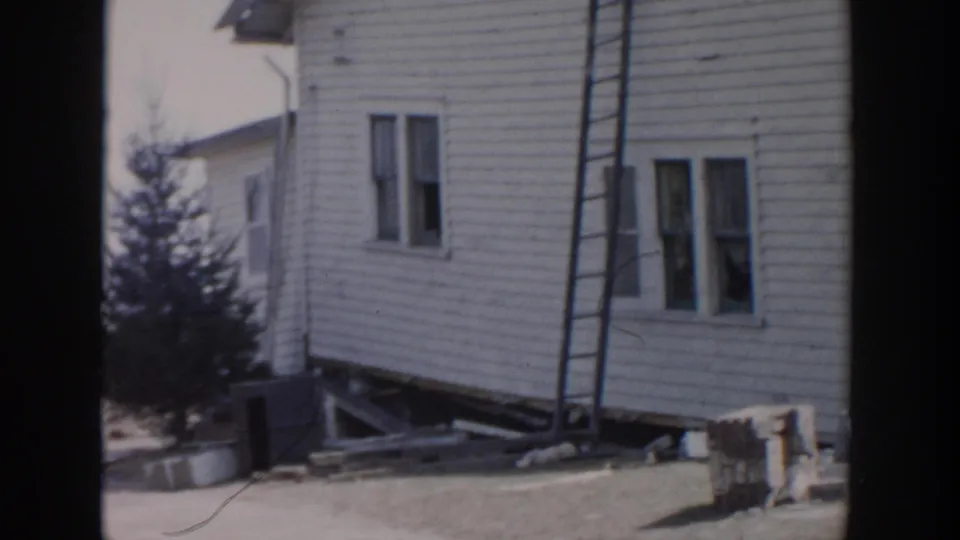 Grand Rapids 1956 frame showing the side of a damaged white house, tilted foundation area, windows, ladder, and sparse yard.