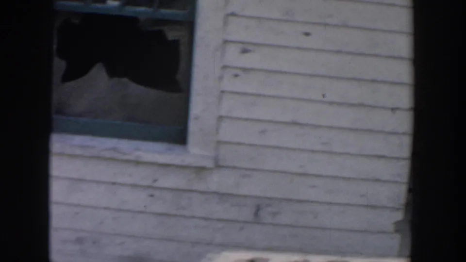 Close 1956 Grand Rapids home-movie frame of a broken window set in white clapboard siding on the damaged house.