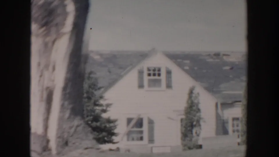 Wider 1956 Grand Rapids view of a small damaged house with missing roof shingles, windows, shrubs, and a tree trunk.