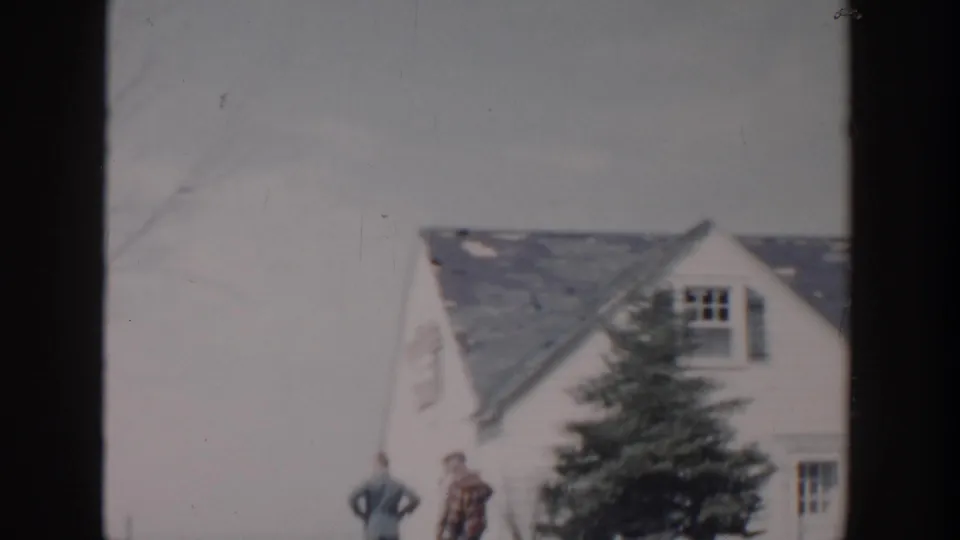 Two men stand outside a damaged Grand Rapids house in 1956, with roof damage and a tree visible nearby.
