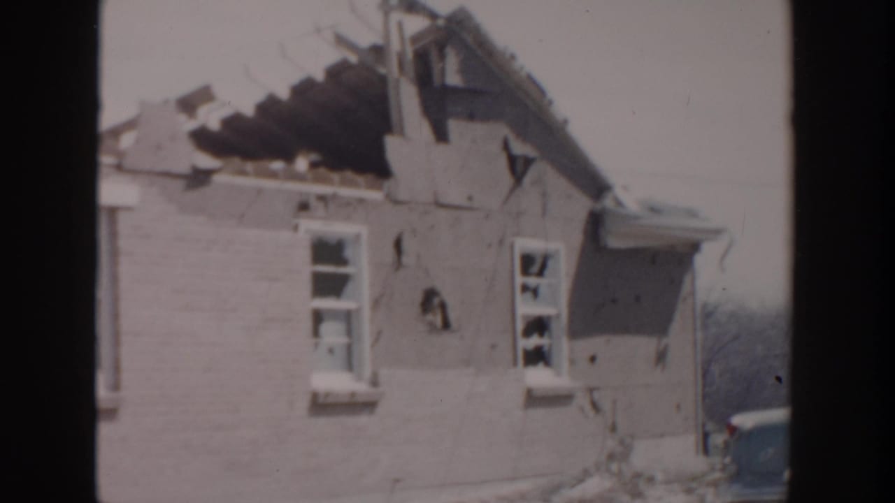 1956 Grand Rapids home-movie frame showing a brick house with severe roof damage, cracked wall sections, and broken windows.