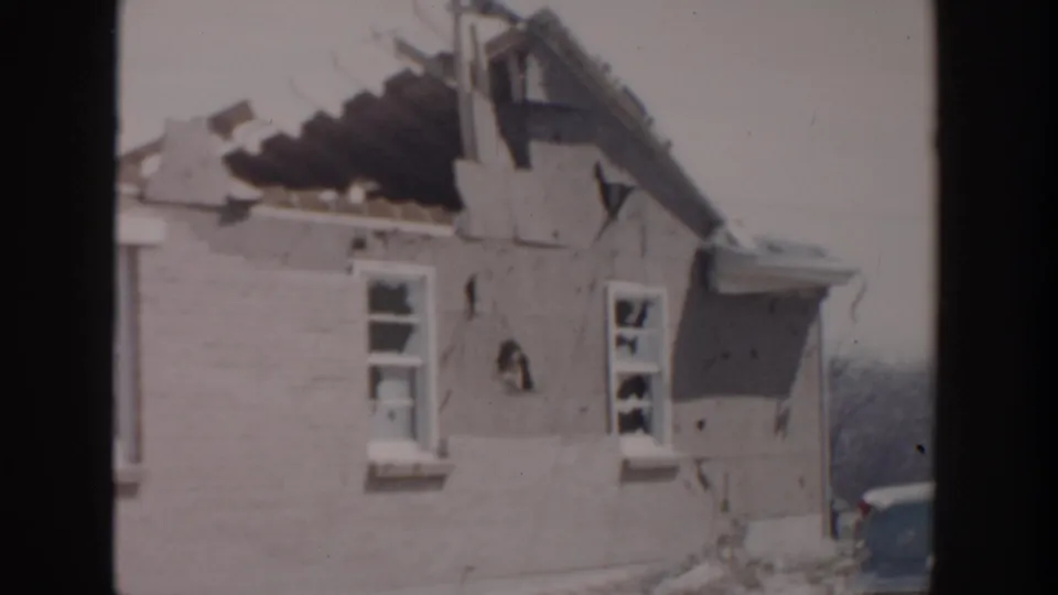 1956 Grand Rapids home-movie frame showing a brick house with severe roof damage, cracked wall sections, and broken windows.