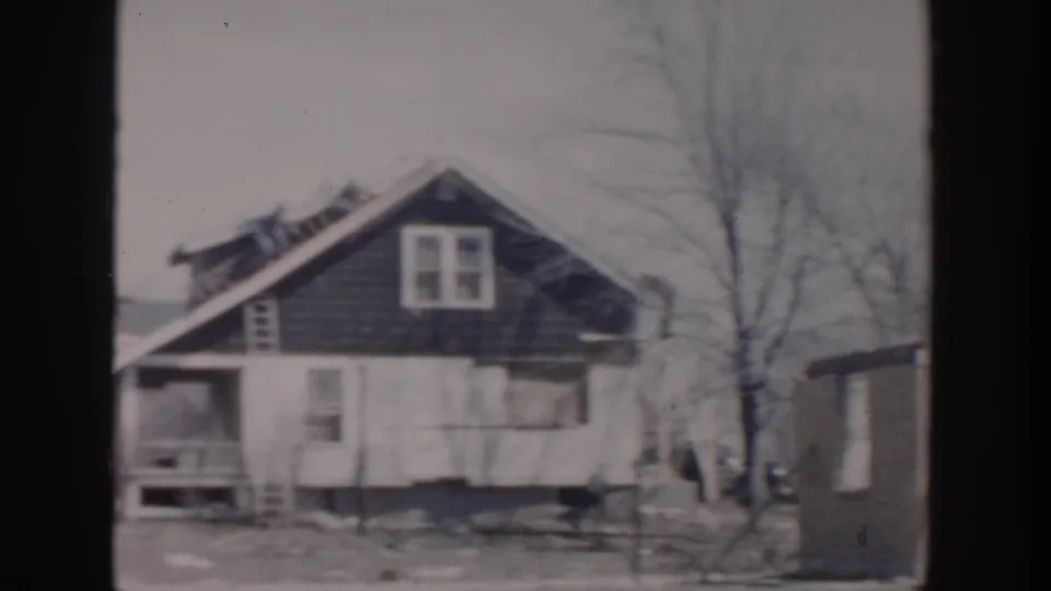 1956 Grand Rapids frame showing a wood-sided house with a damaged roof, bare trees, and debris around the yard.