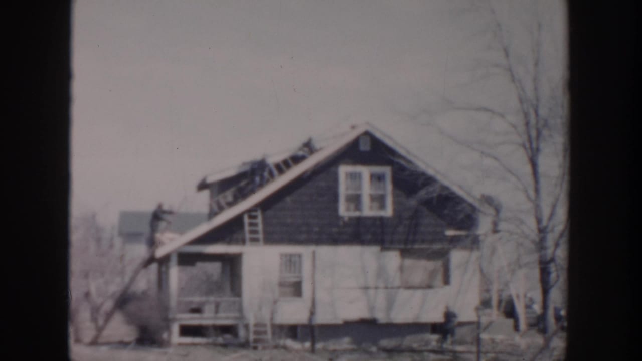 Grand Rapids 1956 home-movie frame showing a weathered two-story house with ladders, workers, bare trees, and a street-facing porch.