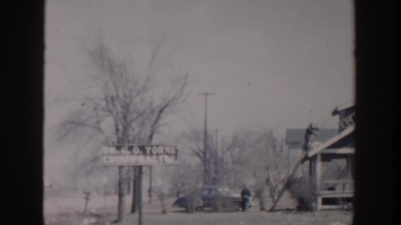 Street view with chiropractor sign, parked car, bare trees, and nearby house.