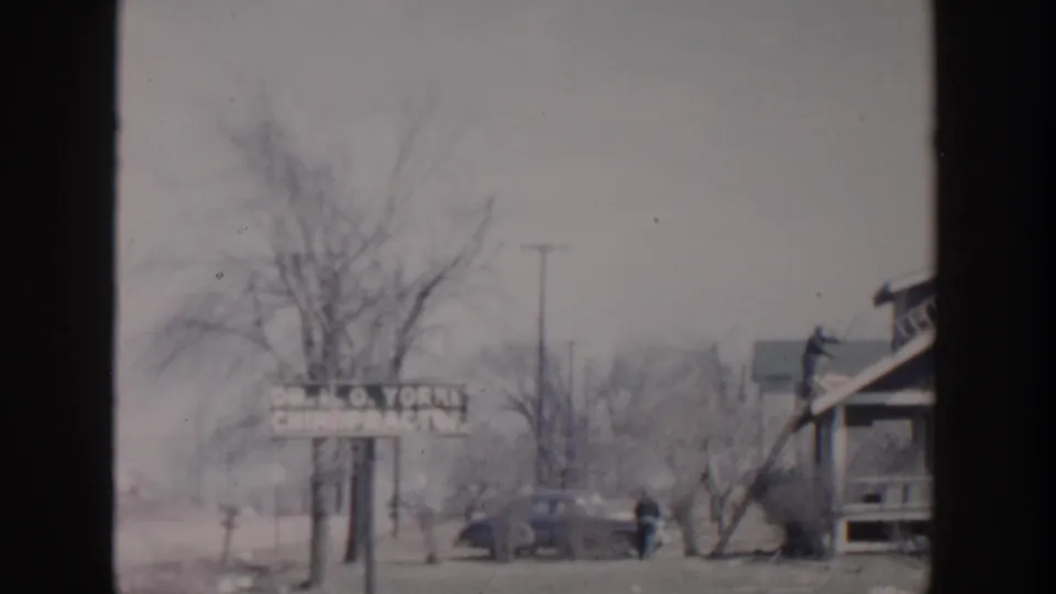 Grand Rapids 1956 street view with a Dr. H. O. Yorks chiropractor sign, parked mid-century car, bare trees, and workers near a house.