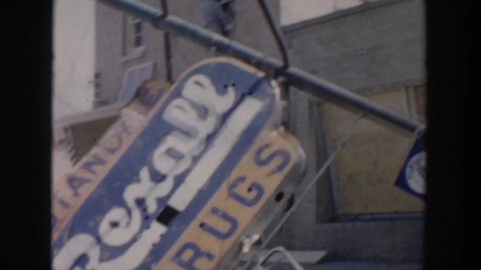 Angled Grand Rapids 1956 frame showing the Rexall Drugs sign close to camera, with a metal bracket, building wall, and partial storefront details.