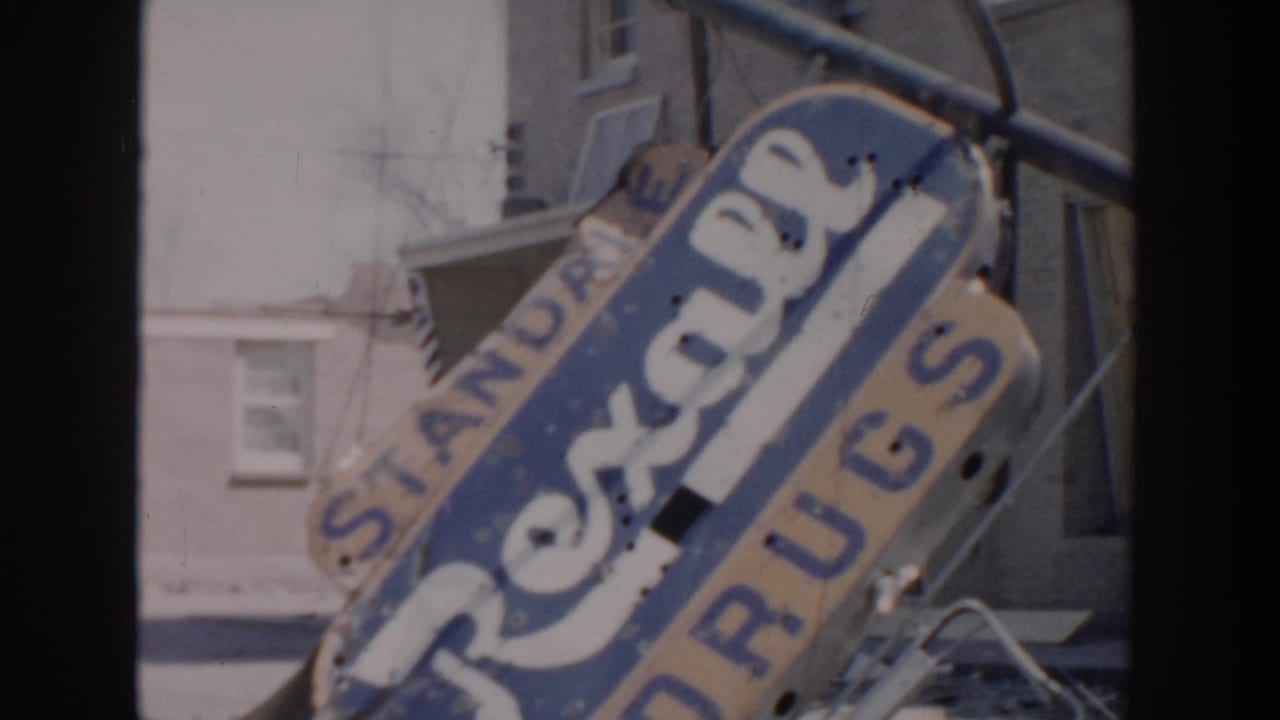 Tilted weathered Rexall Drugs sign beside older houses.