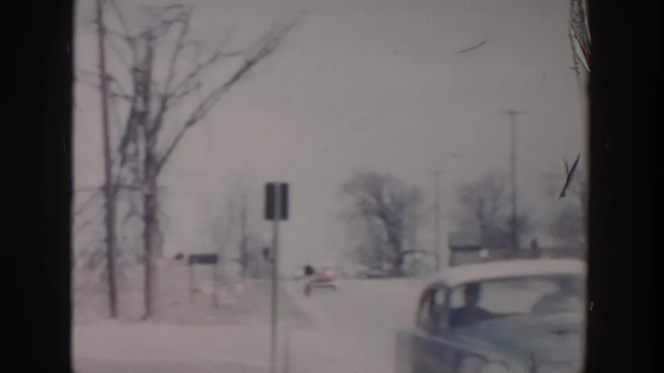 1956 Grand Rapids winter frame with bare trees, roadside signs, and a rounded car partly visible at the right edge.