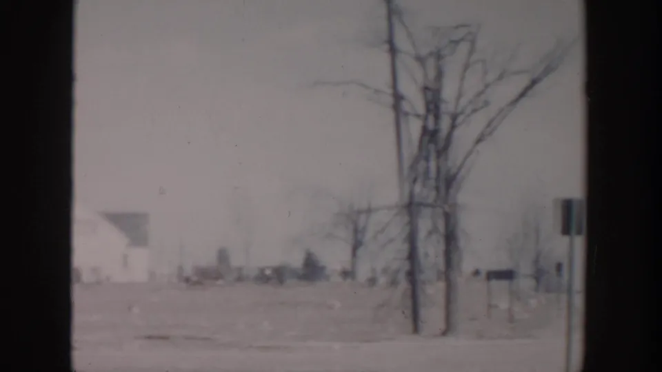 Grand Rapids winter home-movie view showing a snowy roadside, leafless trees, signs, utility poles, and distant buildings.