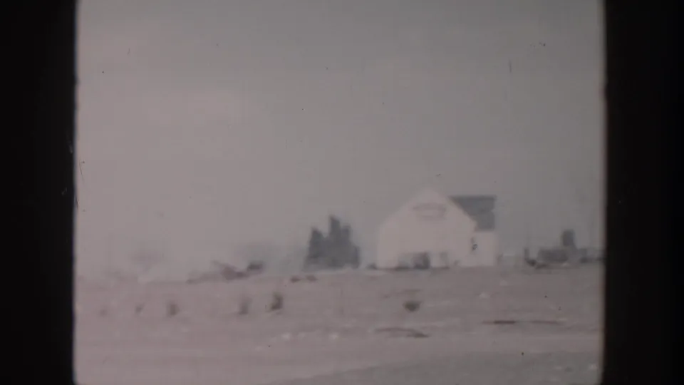 Soft 8mm frame from Grand Rapids showing a distant white building and low houses beyond a flat snowy foreground.