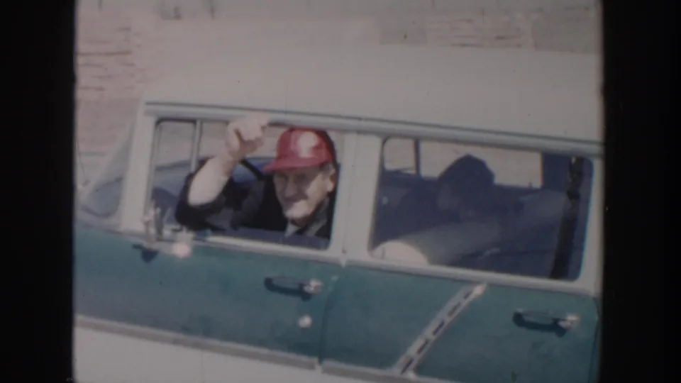 A man in a red cap waves from the open window of a green and white station wagon in 1956 Grand Rapids footage.