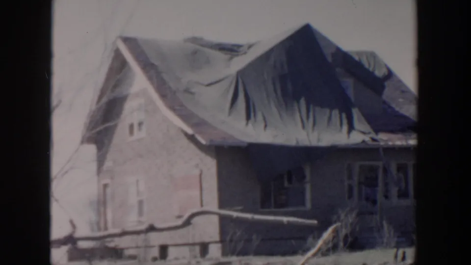 The final frame holds on the brick house exterior, showing the roof tarp, windows, and yard details in soft 8mm color.