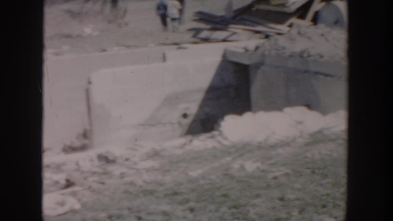1956 Grand Rapids home-movie frame showing concrete work, scattered rubble, lumber, and a small distant figure on a construction site.