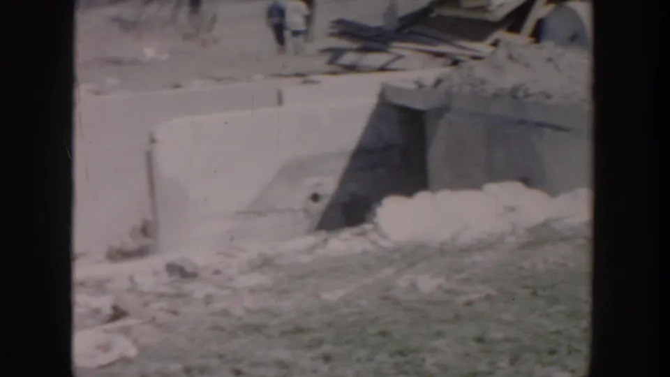 1956 Grand Rapids home-movie frame showing concrete work, scattered rubble, lumber, and a small distant figure on a construction site.