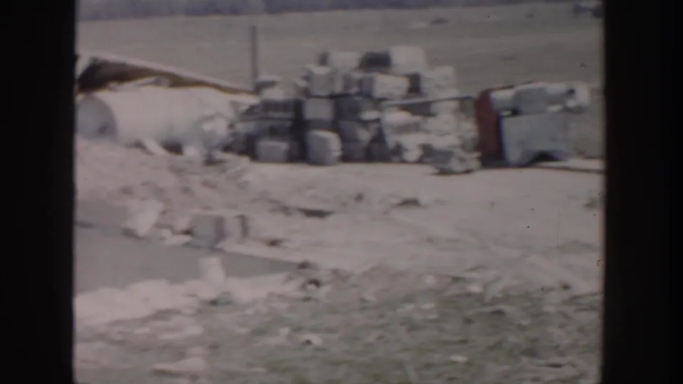 1956 Grand Rapids worksite view with stacked blocks, containers, loose materials, dirt, and grass in a soft 8mm frame.