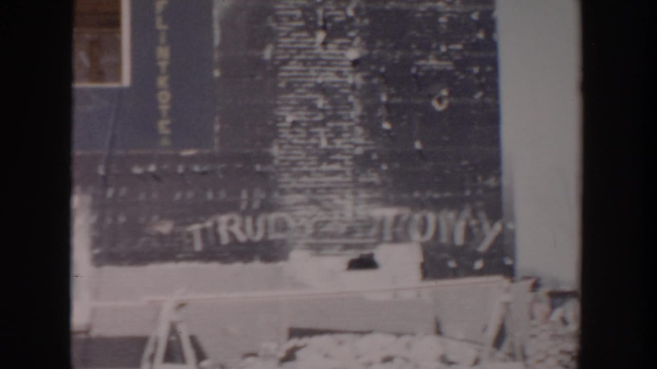 1956 Grand Rapids home-movie frame showing an unfinished house exterior with tarpaper siding, brickwork, a window, and TRUDY TONY graffiti.