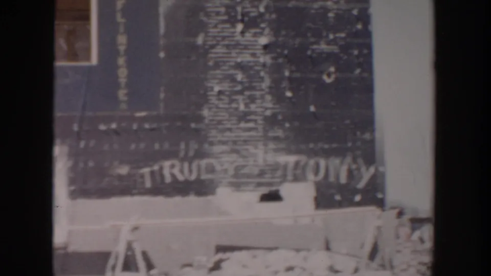 1956 Grand Rapids home-movie frame showing an unfinished house exterior with tarpaper siding, brickwork, a window, and TRUDY TONY graffiti.