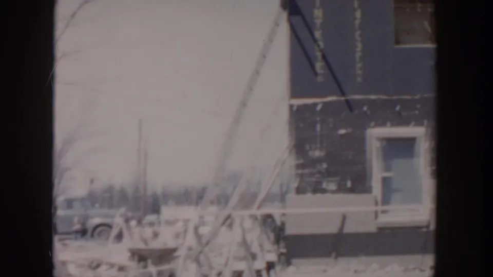 1956 Grand Rapids home-movie view of the unfinished house edge, with tarpaper siding, a window, bare branches, and a parked car nearby.