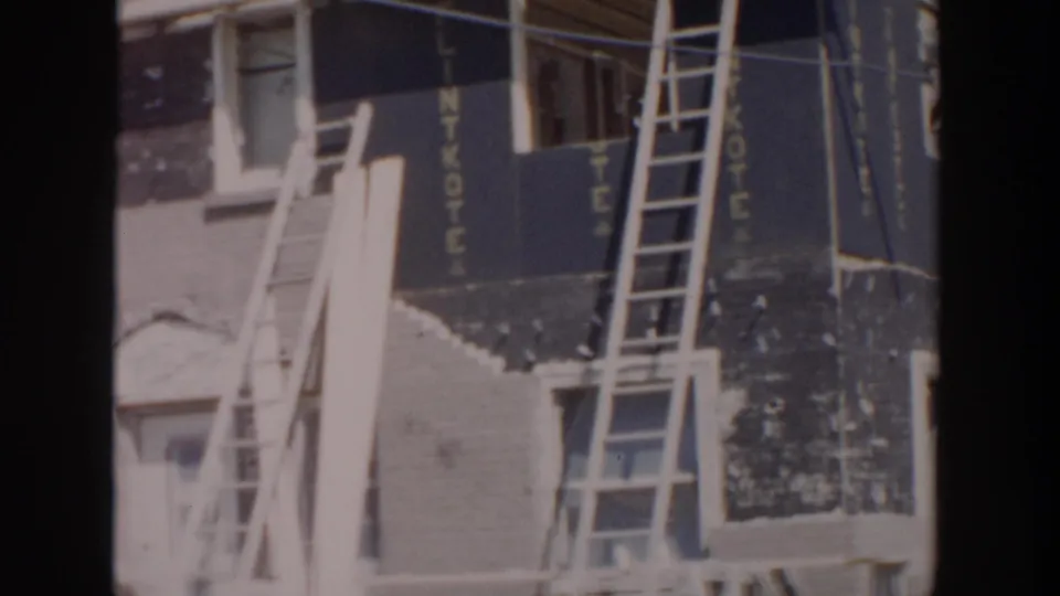 1956 Grand Rapids frame showing ladders against an unfinished house corner, with tarpaper siding, a doorway, and vertical TONY lettering.