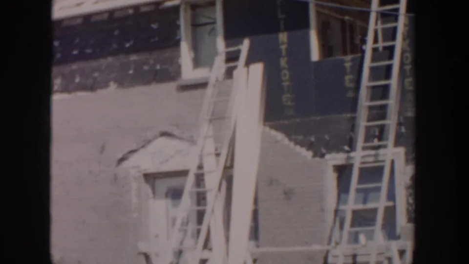1956 Grand Rapids home-movie frame showing ladders against an unfinished residential exterior with dark sheathing and wall surface visible.