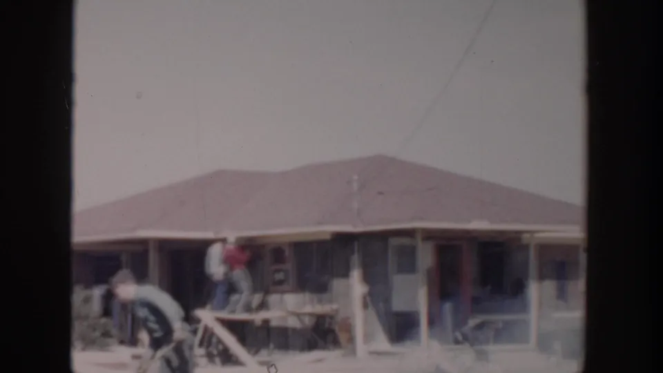 1956 Grand Rapids frame showing workers at a residential construction site beside scaffolding, porch posts, and the front of a single-story house.