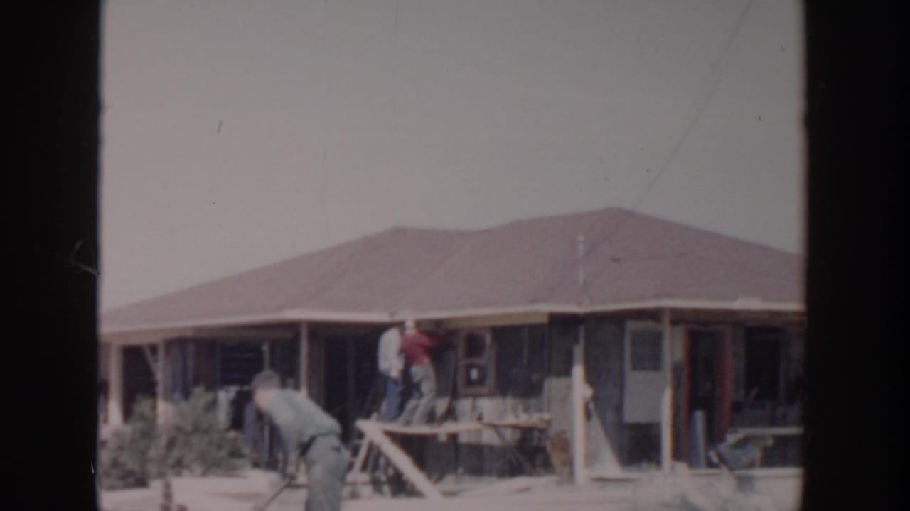 1956 8mm home-movie frame from Grand Rapids showing men remodeling or repairing the exterior of a single-story house.