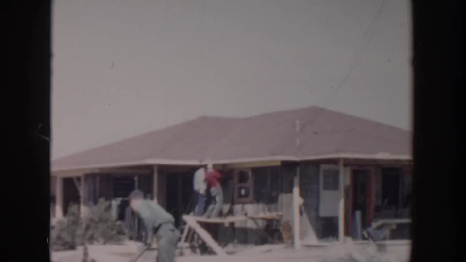 1956 8mm home-movie frame from Grand Rapids showing men remodeling or repairing the exterior of a single-story house.