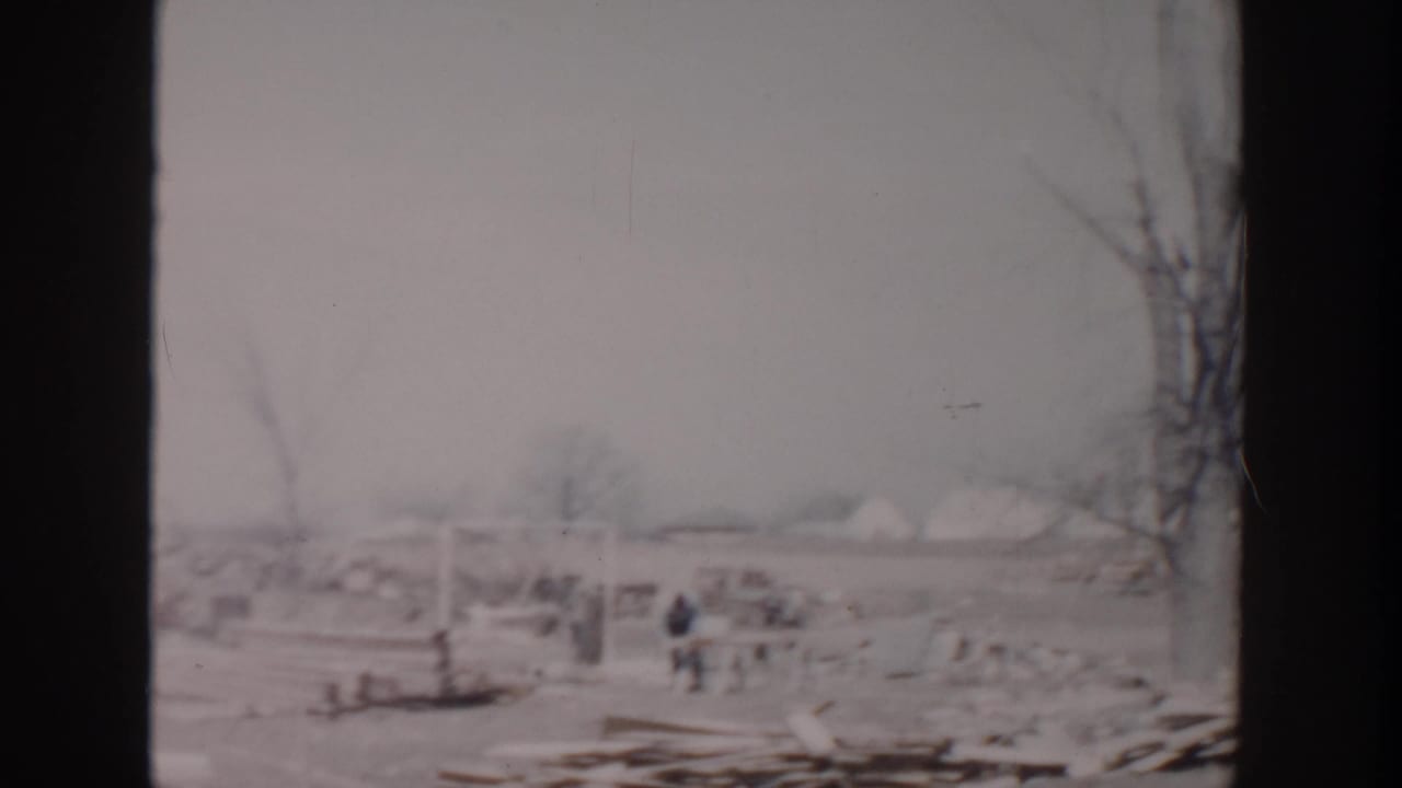1956 Grand Rapids home-movie frame showing a debris-strewn open field, bare trees, and small distant figures moving through wreckage.