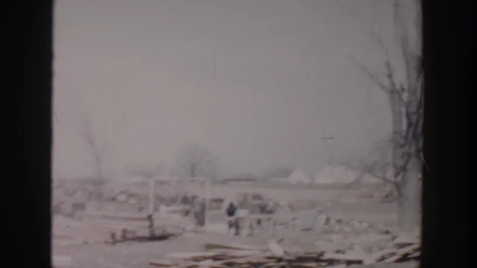 1956 Grand Rapids home-movie frame showing a debris-strewn open field, bare trees, and small distant figures moving through wreckage.