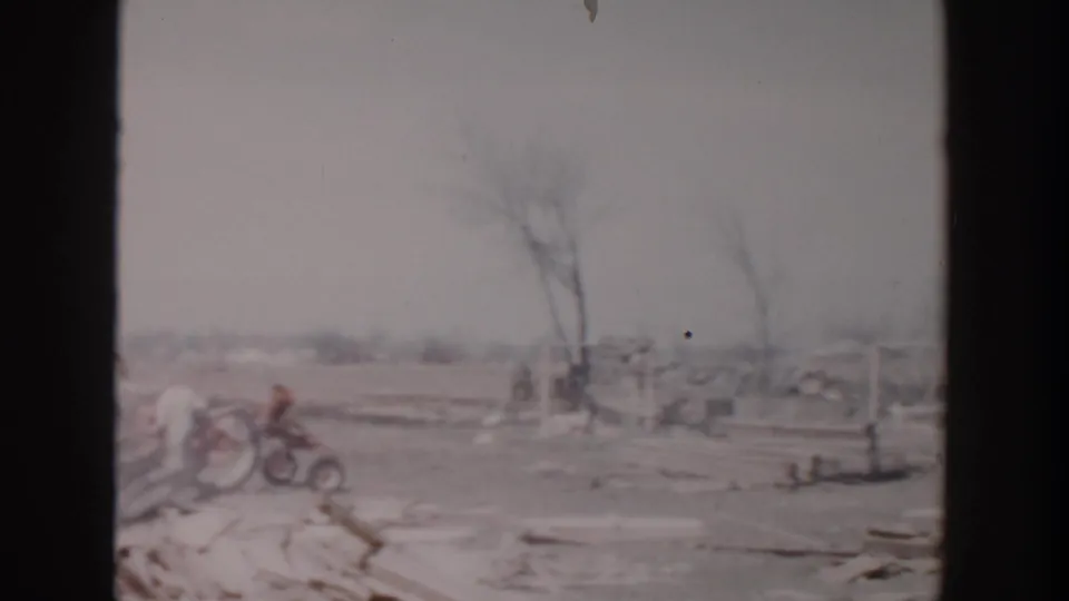 1956 Grand Rapids frame with broken material across an open field and a small tractor or machine near bare trees in the distance.