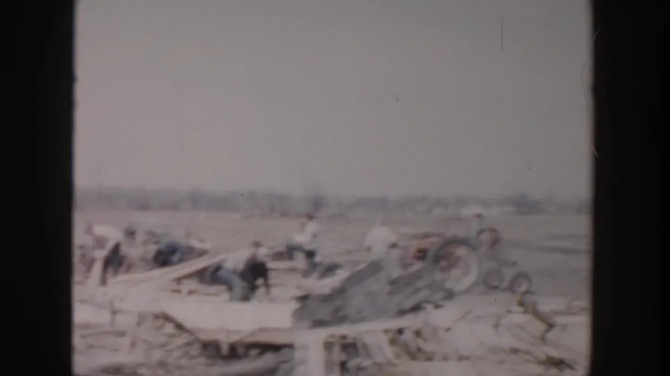 1956 Grand Rapids home-movie view of distant people and a small tractor among broken boards and scattered field debris.