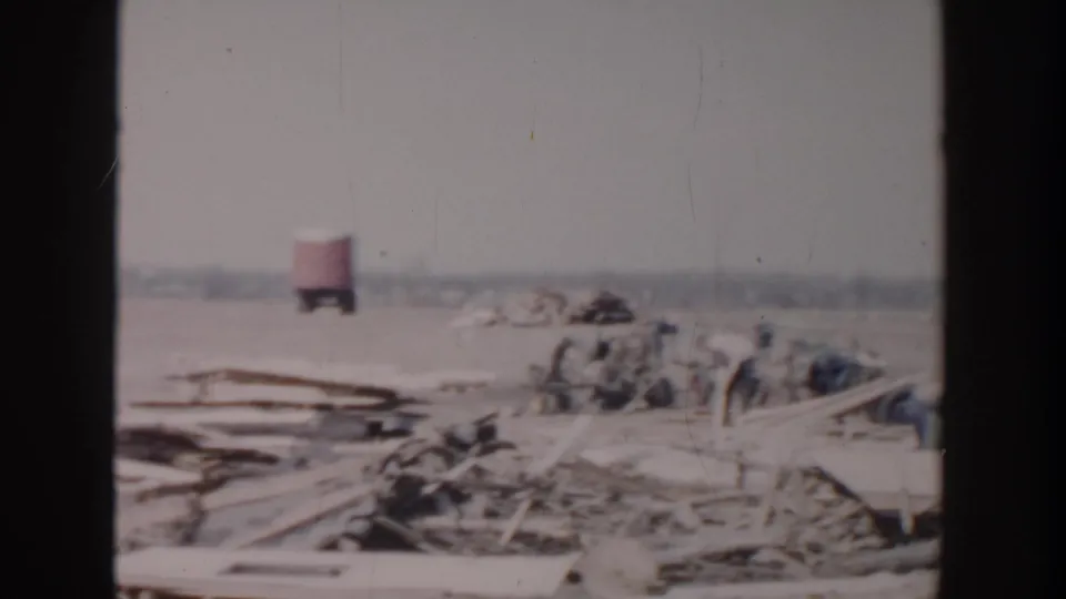 1956 Grand Rapids frame showing a red truck or trailer, clustered distant people, and debris piles across an open field.