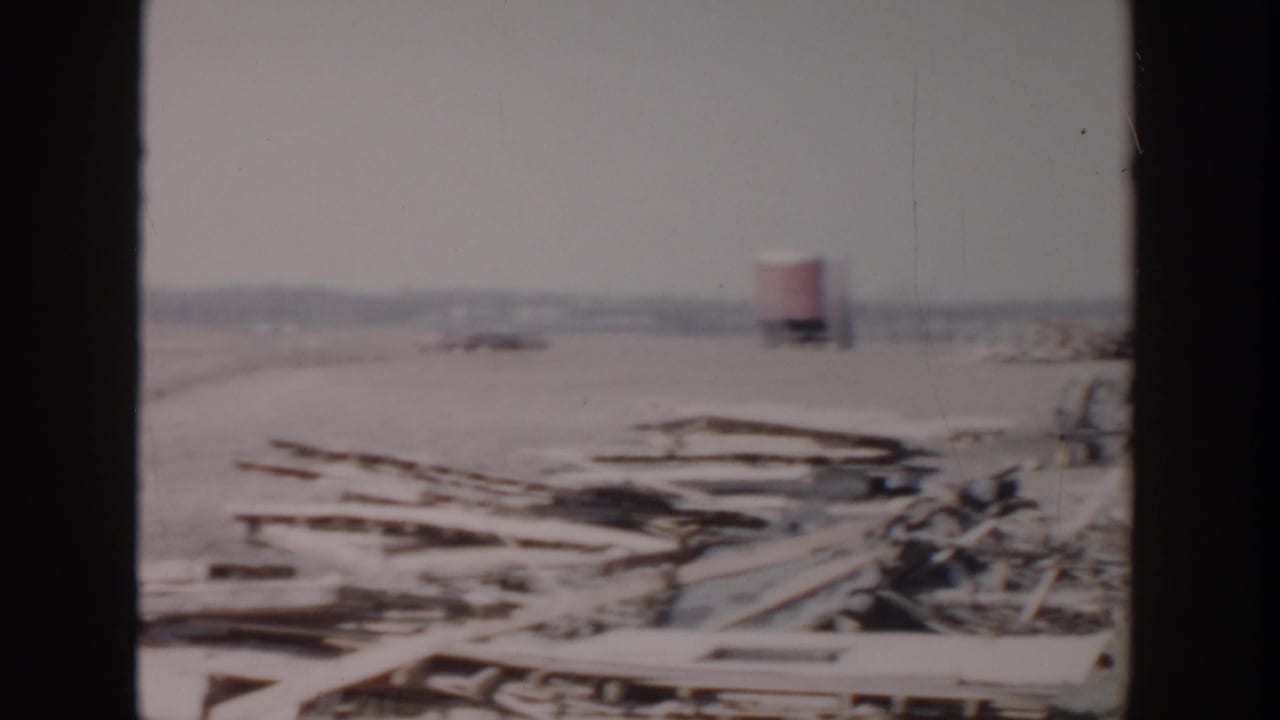 1956 Grand Rapids home-movie frame showing an open lumberyard or construction lot with timber piles and a distant red truck.