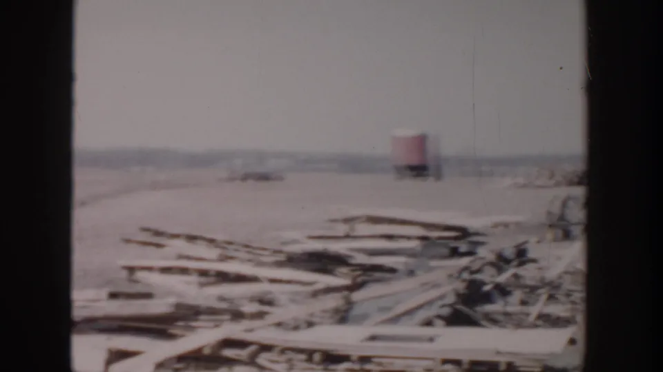 1956 Grand Rapids home-movie frame showing an open lumberyard or construction lot with timber piles and a distant red truck.