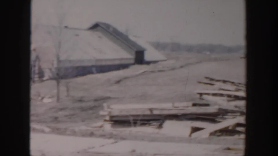 1956 Grand Rapids frame with a small yard building, bare trees, scattered lumber, and open ground under a muted sky.