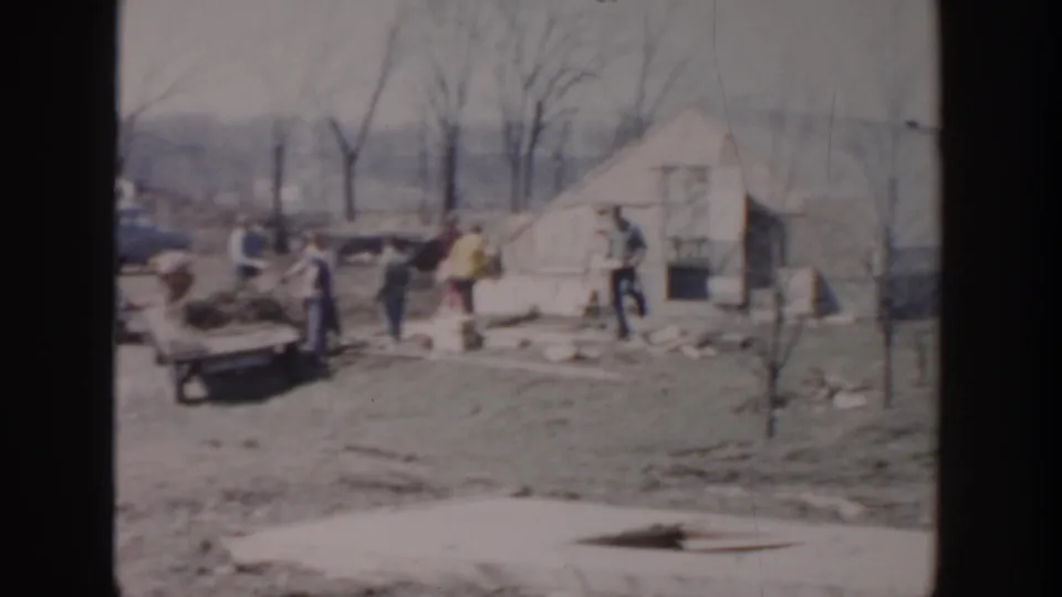 1956 Grand Rapids home-movie view of workers near a small building, with boards, yard debris, and bare trees around them.