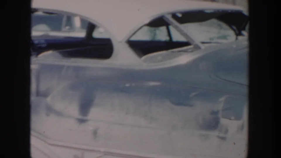 Close 1956 Grand Rapids home-movie frame showing the side of a light-colored period car with another dark vehicle partly behind it.