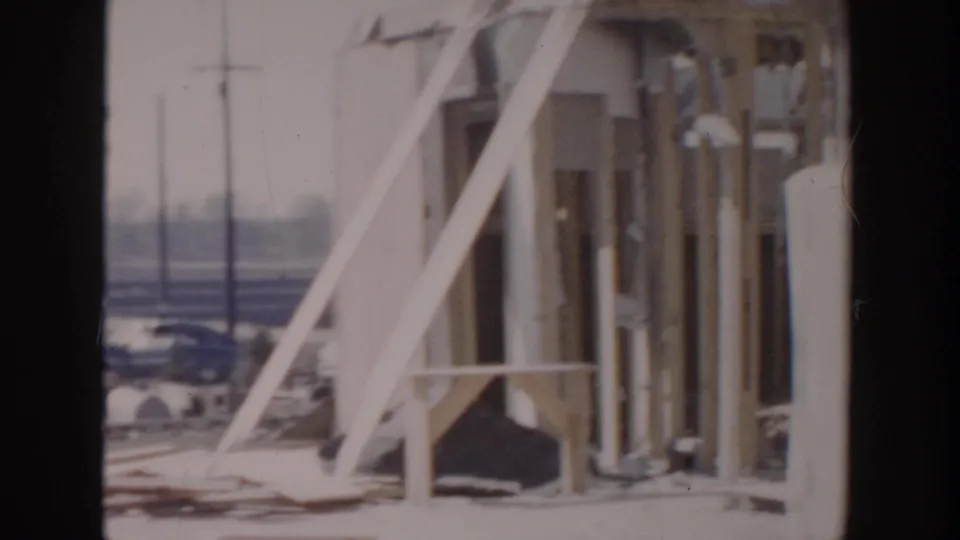 1956 Grand Rapids frame showing exposed wooden framing, pipes, diagonal supports, and parked cars beyond the construction site.