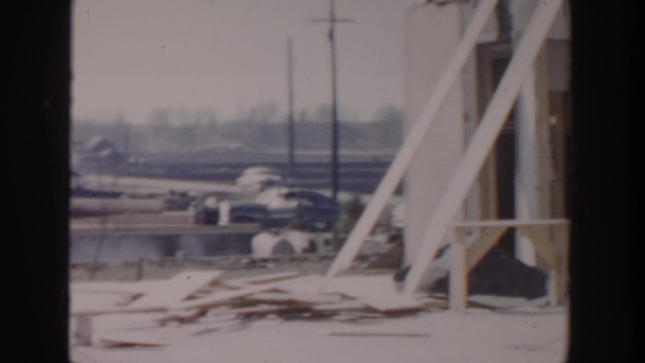 White structural framing and lumber near a roadside worksite with cars and utility poles in the background.