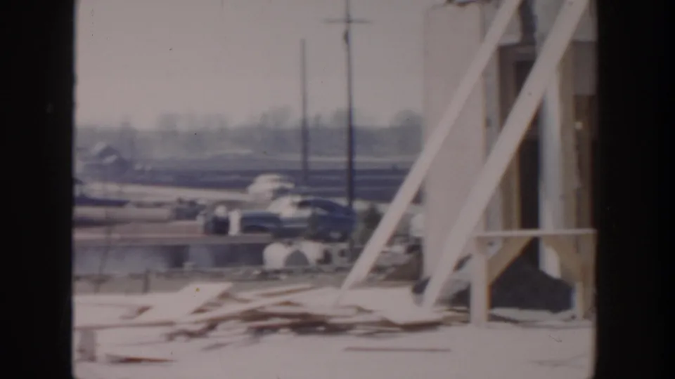 1956 Grand Rapids home-movie frame showing white framing, scattered lumber, cars, and utility poles around a construction site.